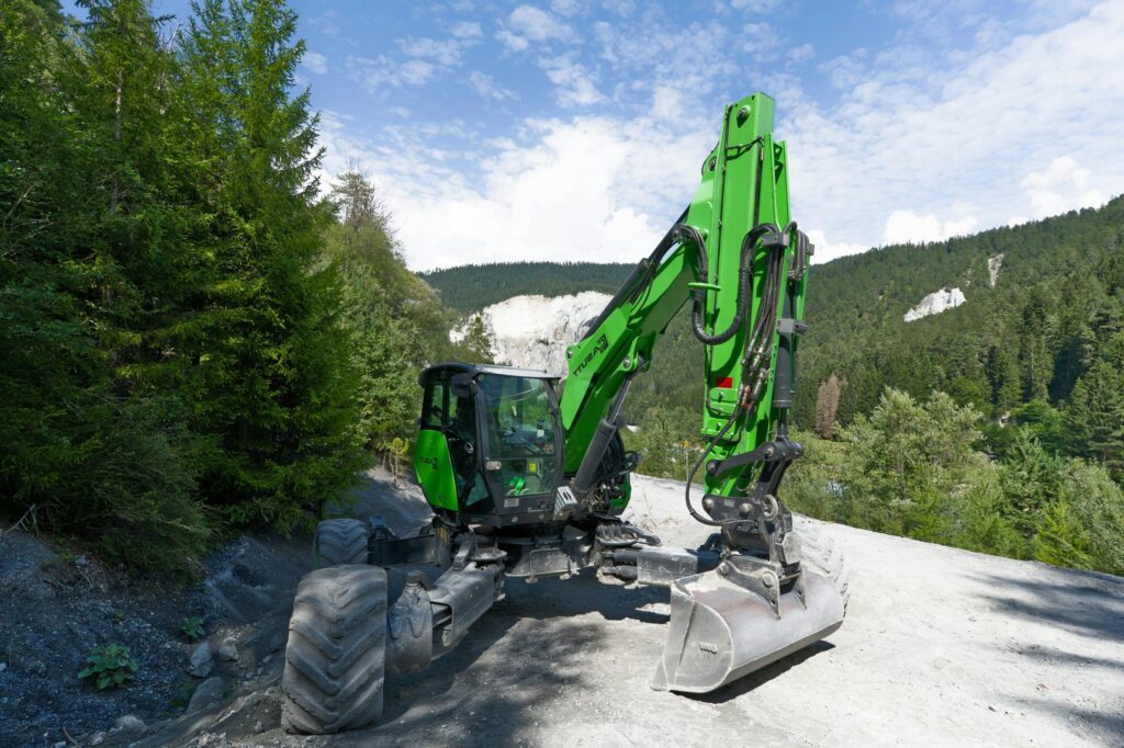 pexels-photo-32555907-32555907 A green excavator operating in a scenic Swiss alpine forest with clear skies.